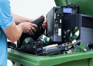 Laptops, computers, motherboards, game consoles, cameras & other electronic waste being placed inside a green e-waste recycling bin for disposal.